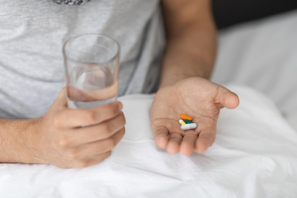 A man is relaxing in bed holding a glass of water in one hand and a selection of pills in the other